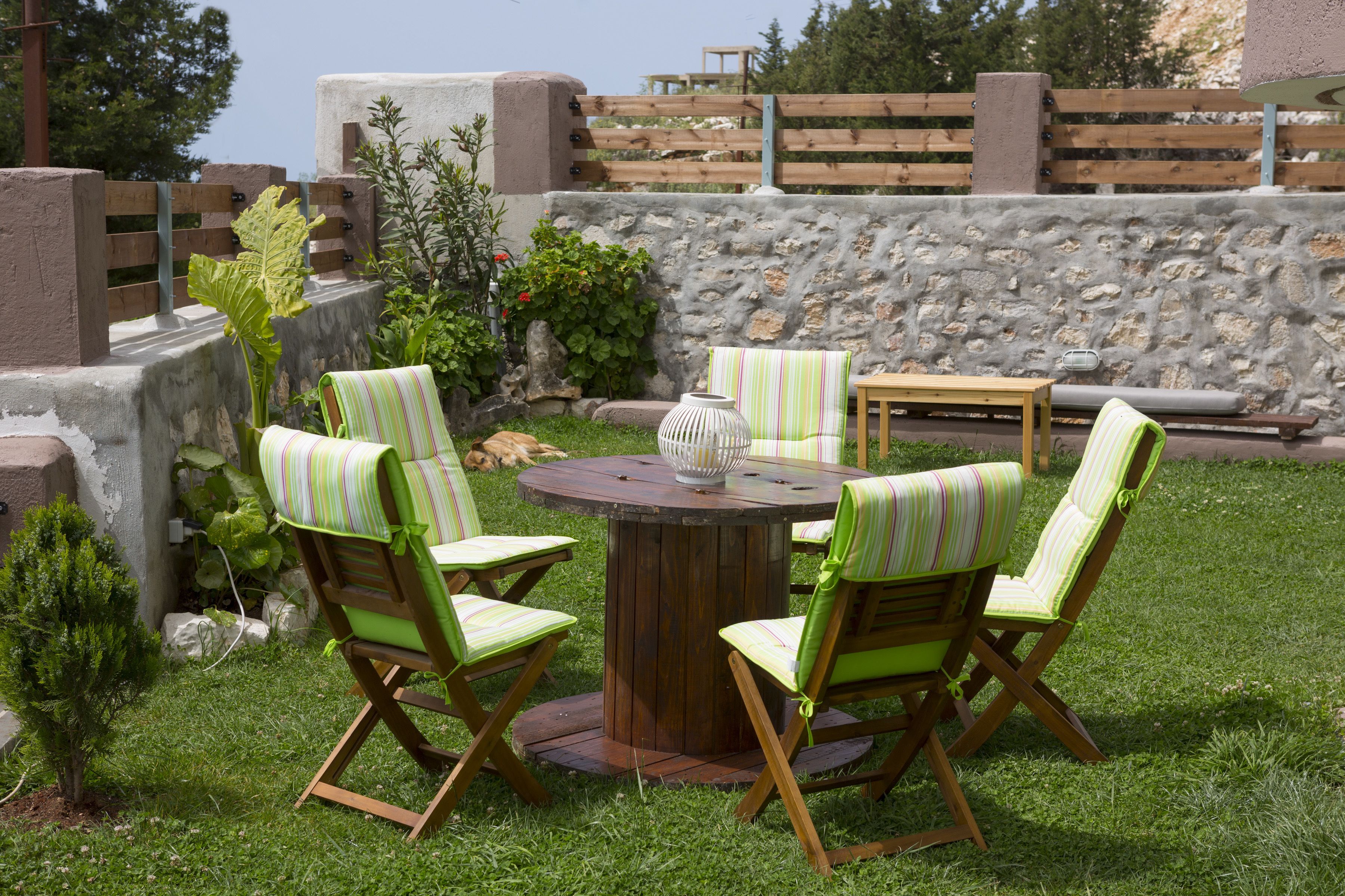 Un patio al aire libre con una mesa redonda de madera rodeada por seis sillas a rayas, una mesa pequeña redonda con una lámpara y un muro de piedra al fondo.