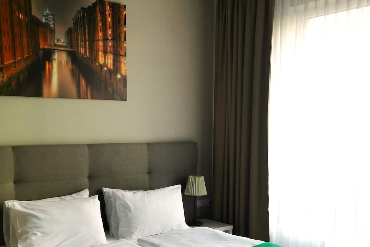 A neatly made bed with white linens and a green throw, a nightstand with a lamp, and a painting of a cityscape on the wall.