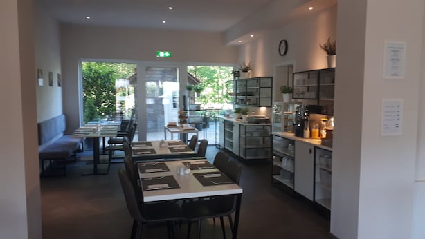 A restaurant with a long counter, tables, and chairs. There is a kitchen area with shelves and appliances on the right. - Hotel Gutsmühle (Obernburg)