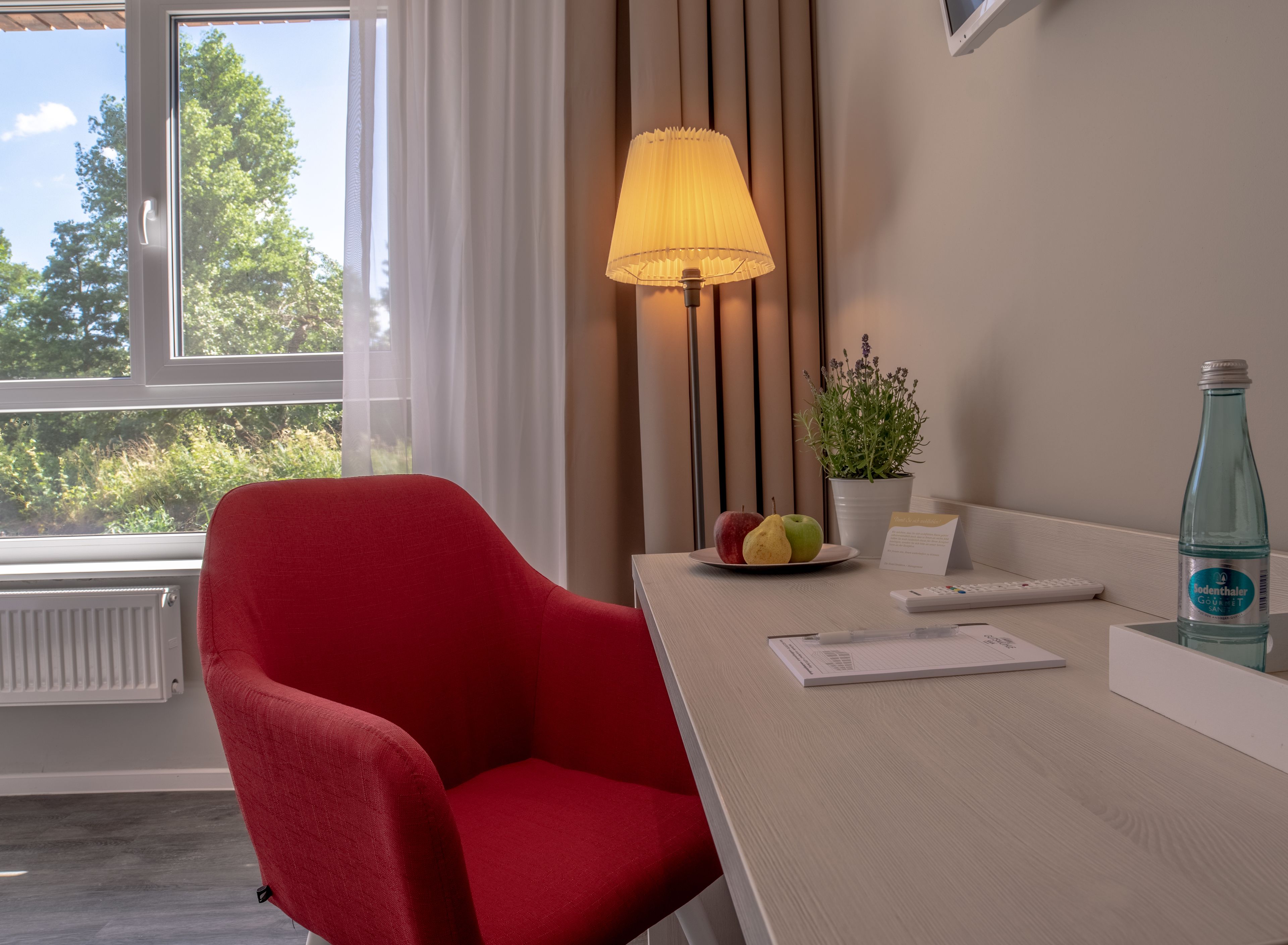 Et hotelværelse med en rød stol, et bord med en lampe, en tallerken med frugt, en flaske og en potteplante.