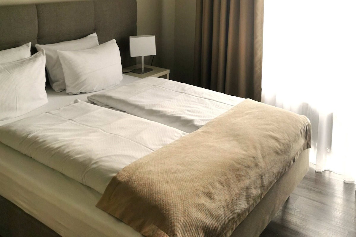 A neatly made bed with white linens, a grey headboard, and a brown throw blanket. There is a bedside table with a lamp and a curtain partially drawn to the side.