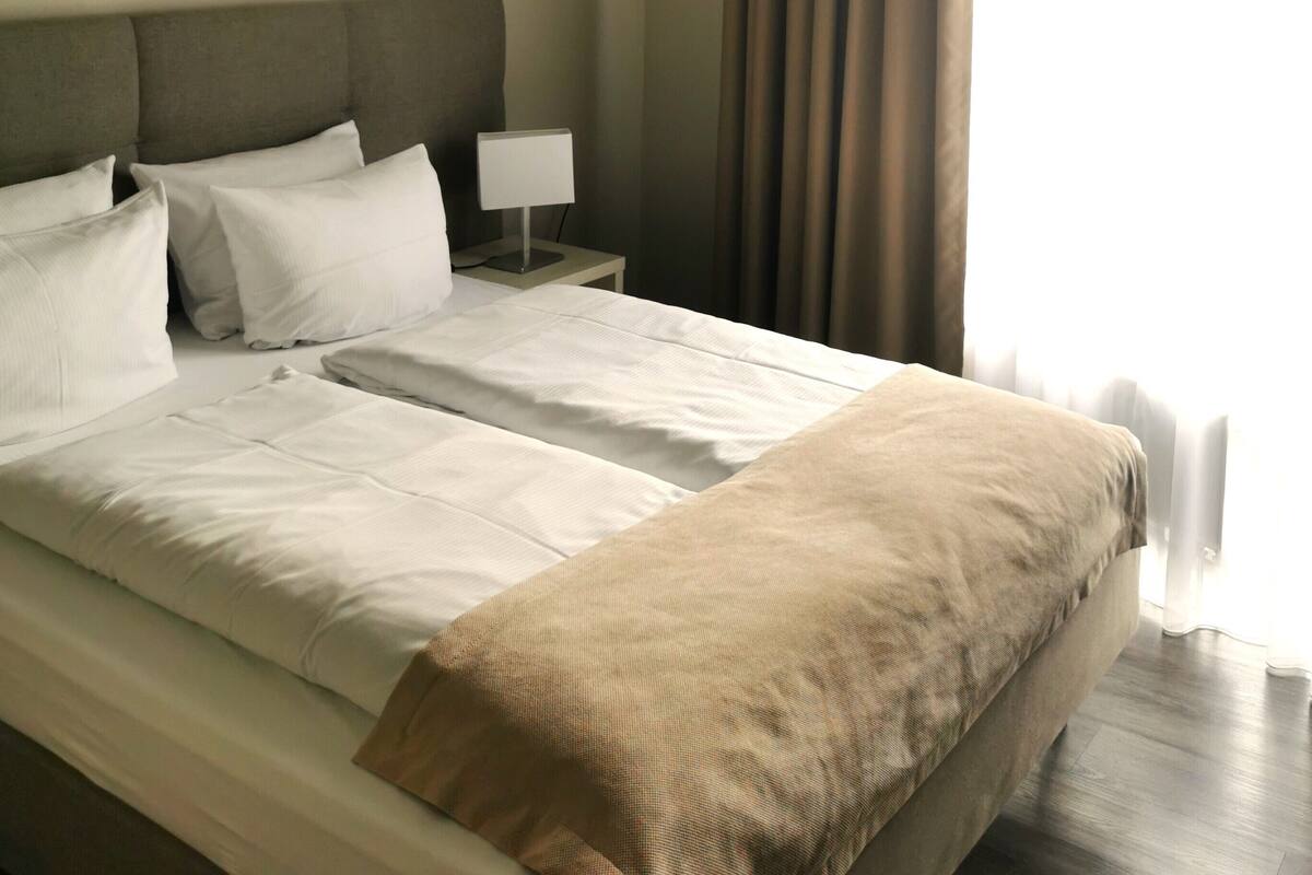 A neatly made bed with white linens and a beige throw blanket, a bedside table with a lamp, and a window with curtains.