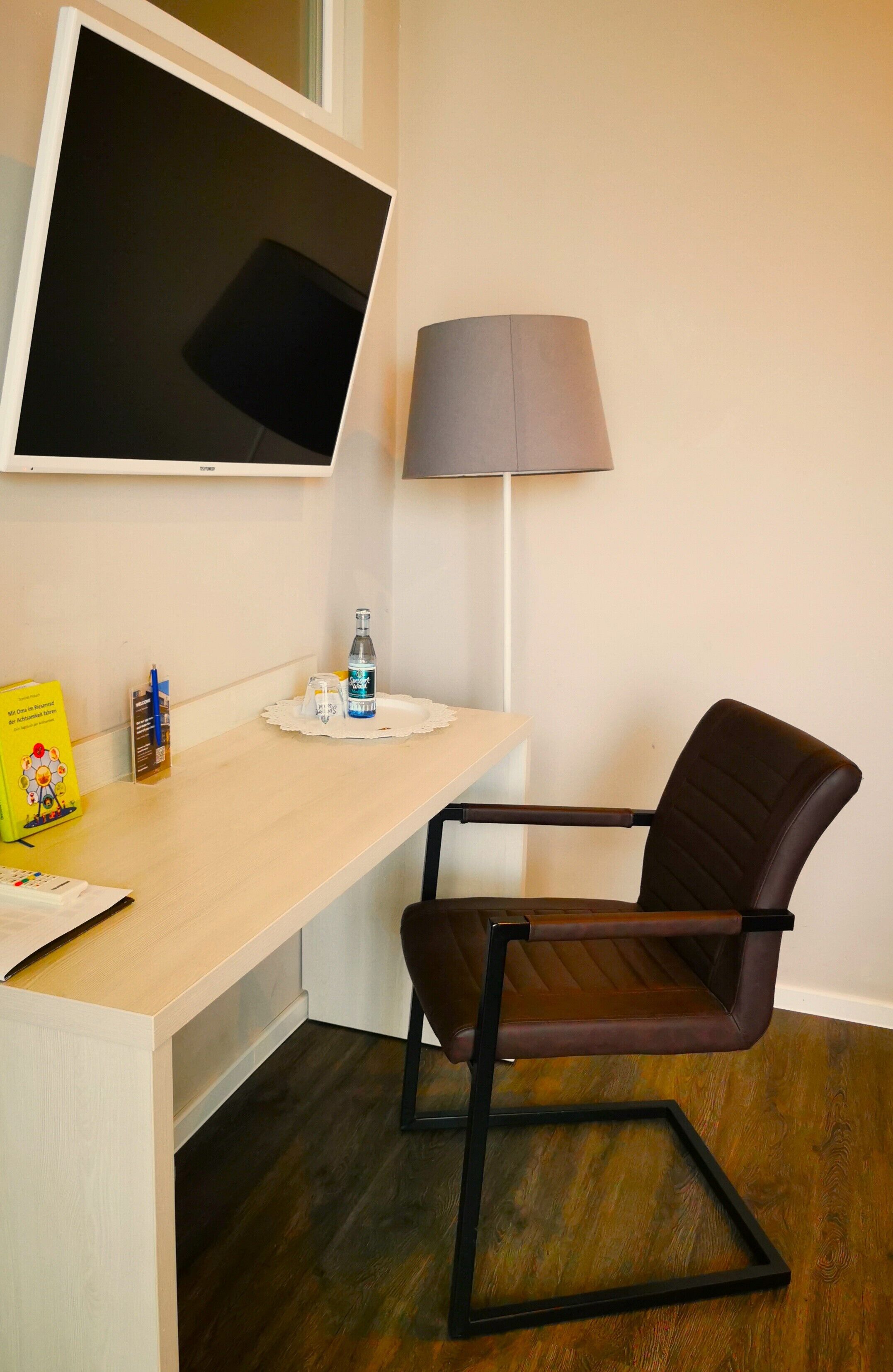 A room with a desk, a chair, a TV mounted on the wall, a lamp, and a bottle of water.