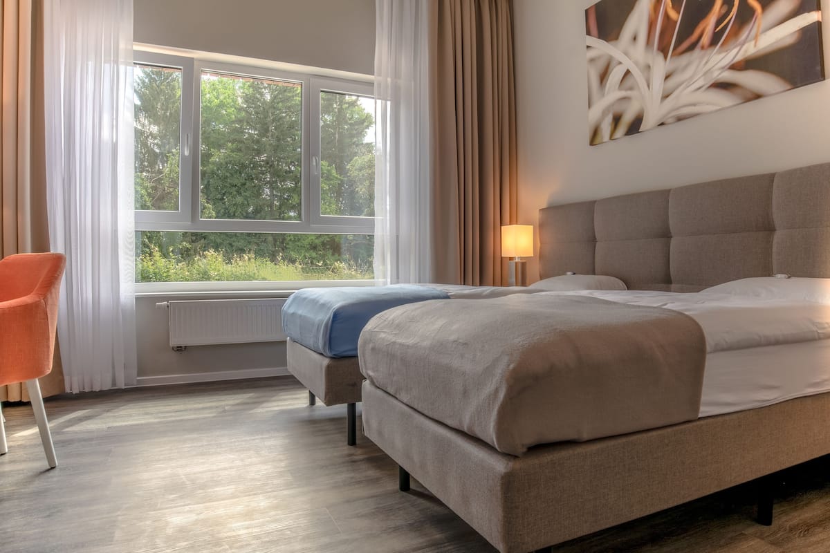 A bedroom with a bed, a chair, a window with curtains, a radiator, and a wall-mounted lamp.