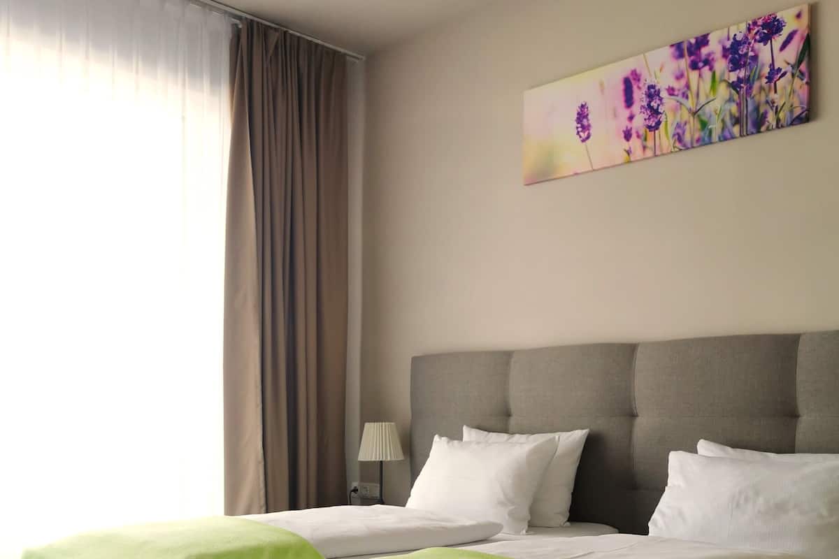 A double bed with a grey headboard and two green pillows, a bedside table with a lamp, and a painting on the wall.