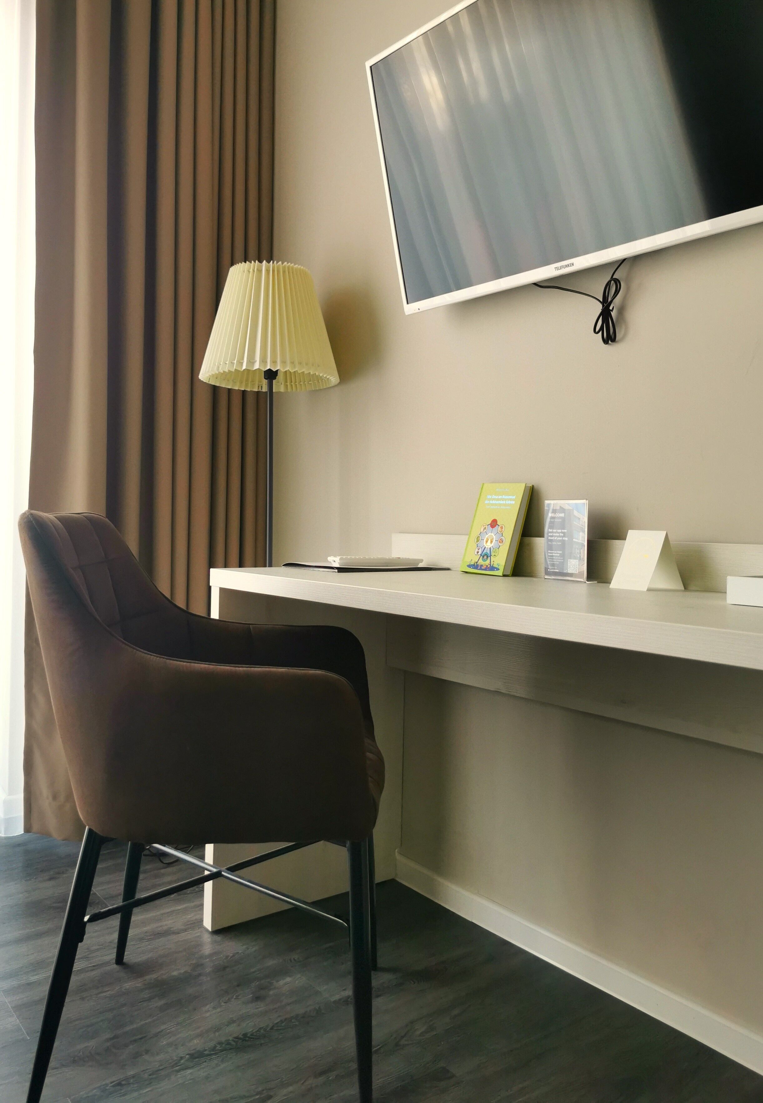 A hotel room with a desk, a chair, a flat-screen TV mounted on the wall, a lamp, and a window with curtains.