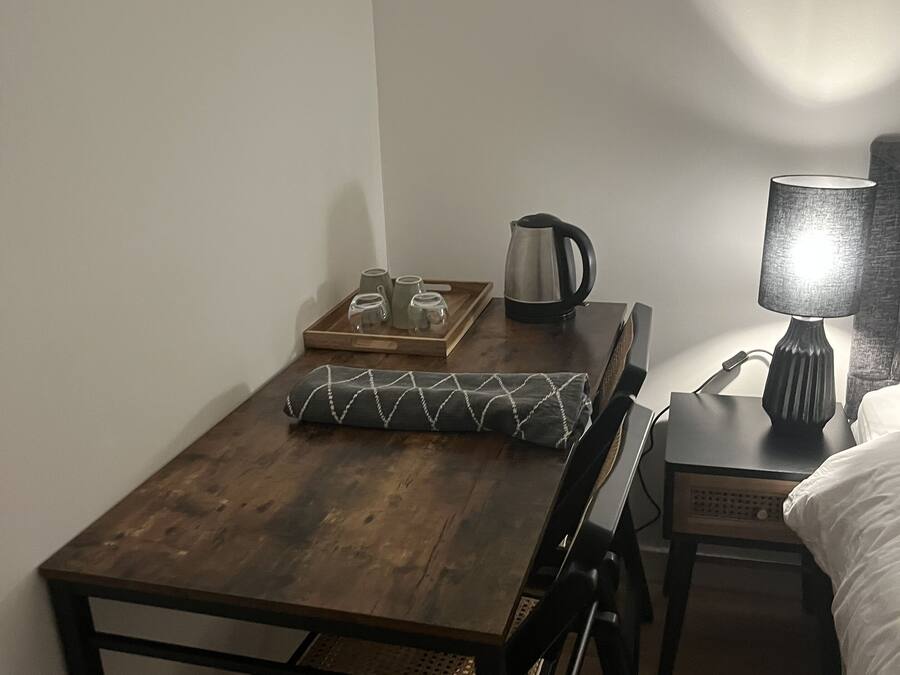 Una habitación pequeña y acogedora con una mesa de madera, una silla, una cama, una mesita de noche y una lámpara.