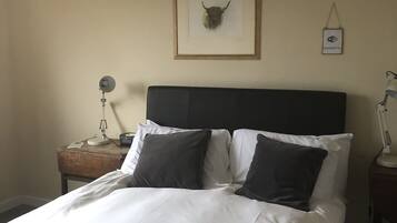 A neatly made bed with white and dark pillows, a framed animal head on the wall, a bedside lamp, and a small table with a lamp and a phone.