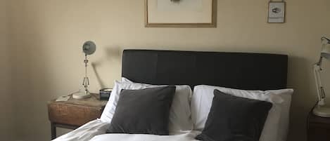 A neatly made bed with white and dark pillows, a framed animal head on the wall, a bedside lamp, and a small table with a lamp and a phone.