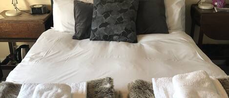 Una cama bien hecha con almohadas blancas y grises, un cabecero oscuro y una mesita de noche con una lámpara y una caja de pañuelos.