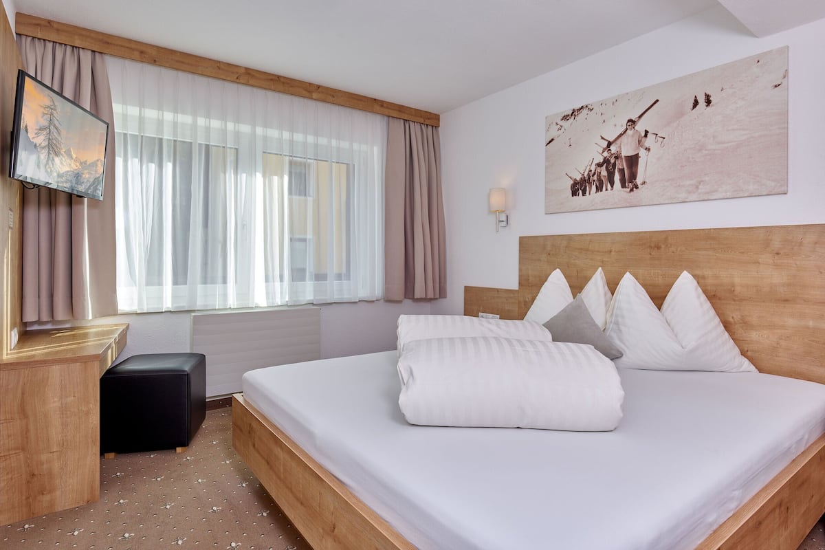 A hotel room with a large bed, a wooden headboard, a painting on the wall, and a window with sheer curtains.