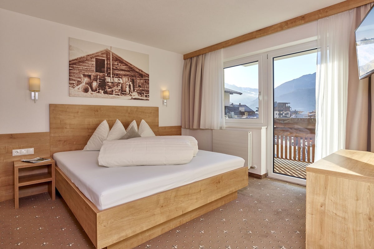 A hotel room with a wooden bed, a nightstand, a wall-mounted lamp, a window with a view of mountains, and a picture of a wooden house.
