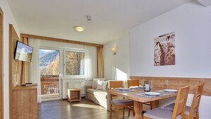 A dining area with a wooden table and chairs, a television mounted on a wooden cabinet, and a window with a view of trees and mountains. - Gästehaus Bergkristall (Soelden)