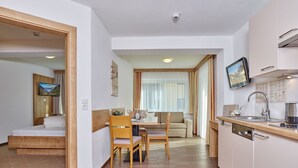 A modern hotel room with a wooden bed, a dining area with a table and chairs, a kitchenette with a sink and stove, and a television mounted above the counter. - Gästehaus Bergkristall (Soelden)