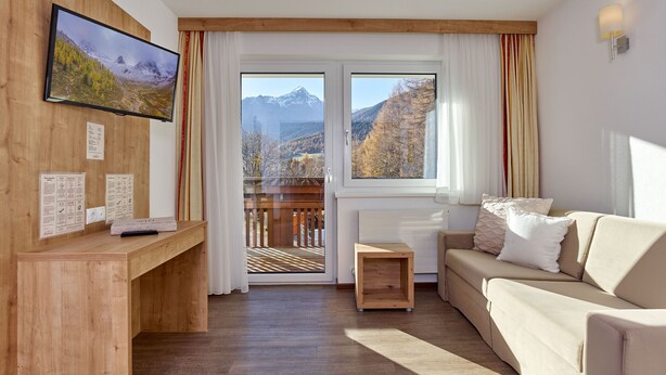 A modern living room with a wooden desk, a large TV, a sofa, and a balcony with a mountain view. - Gästehaus Bergkristall (Soelden)