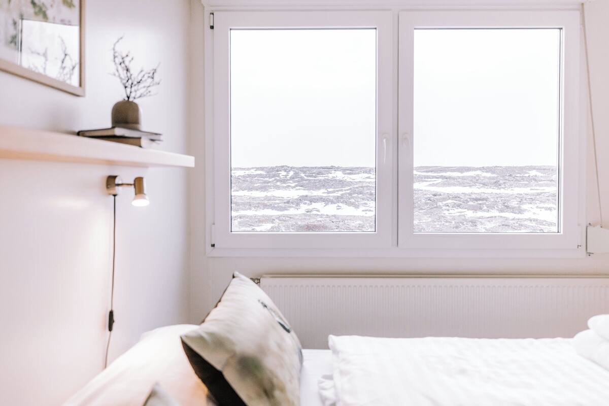 A bedroom with a large window, a bed, a bedside table, and a wall-mounted shelf with a plant.