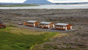 A remote lodge with three wooden cabins, a gravel driveway, and a view of a lake and mountain in the distance. - Hlíð Bed and Breakfast (Myvatn)