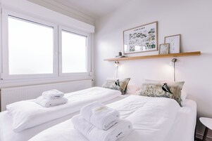 Quarto de hotel com duas camas, roupa de cama branca, janela com persianas, prateleira com molduras e mesa de cabeceira com abajur.