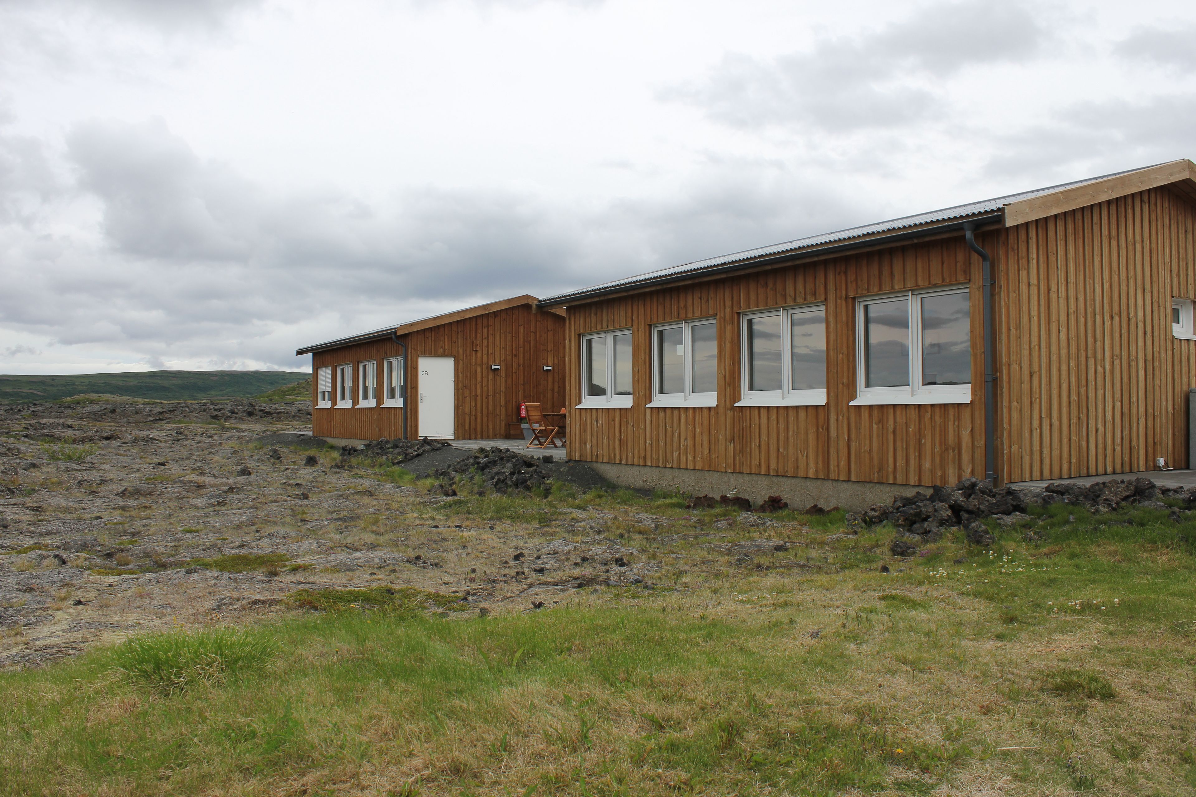 A wooden building with large windows and a door, situated on a rocky terrain with sparse grass.