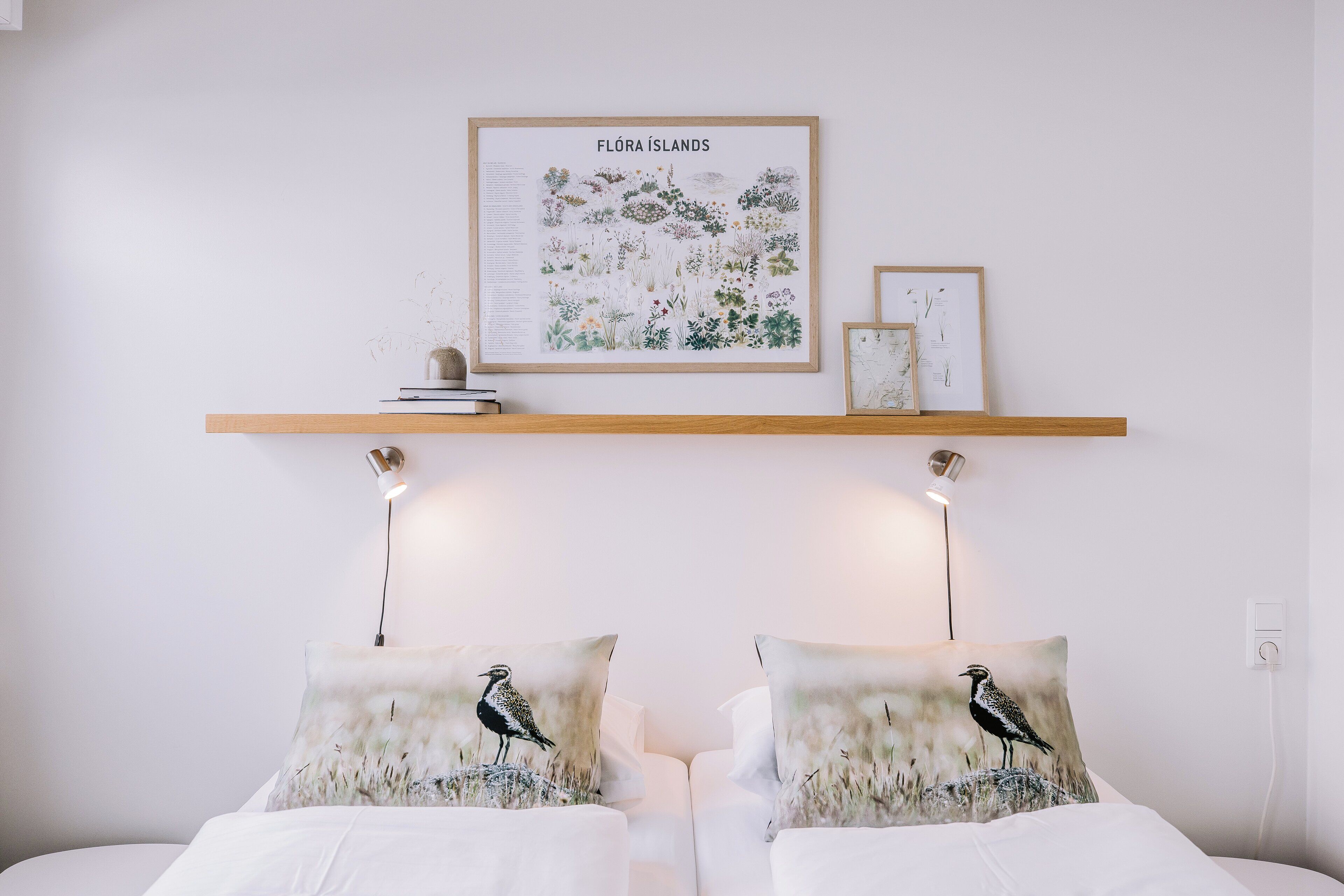 Un dormitorio con una cama, dos almohadas con diseños de pájaros, una estantería de pared con libros y un cuadro enmarcado, e ilustración botánica titulada 'Flóra Islands' en la pared.
