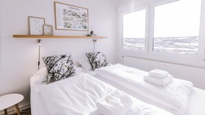 A hotel room with two beds, a window with a view of a snowy landscape, a shelf with framed botanical prints, and a small round table. - Hlíð Bed and Breakfast (Myvatn)