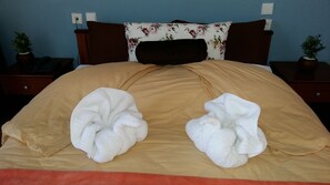 A bed with a beige bedspread, two large white towels, a floral pillow, and two bedside tables with potted plants.