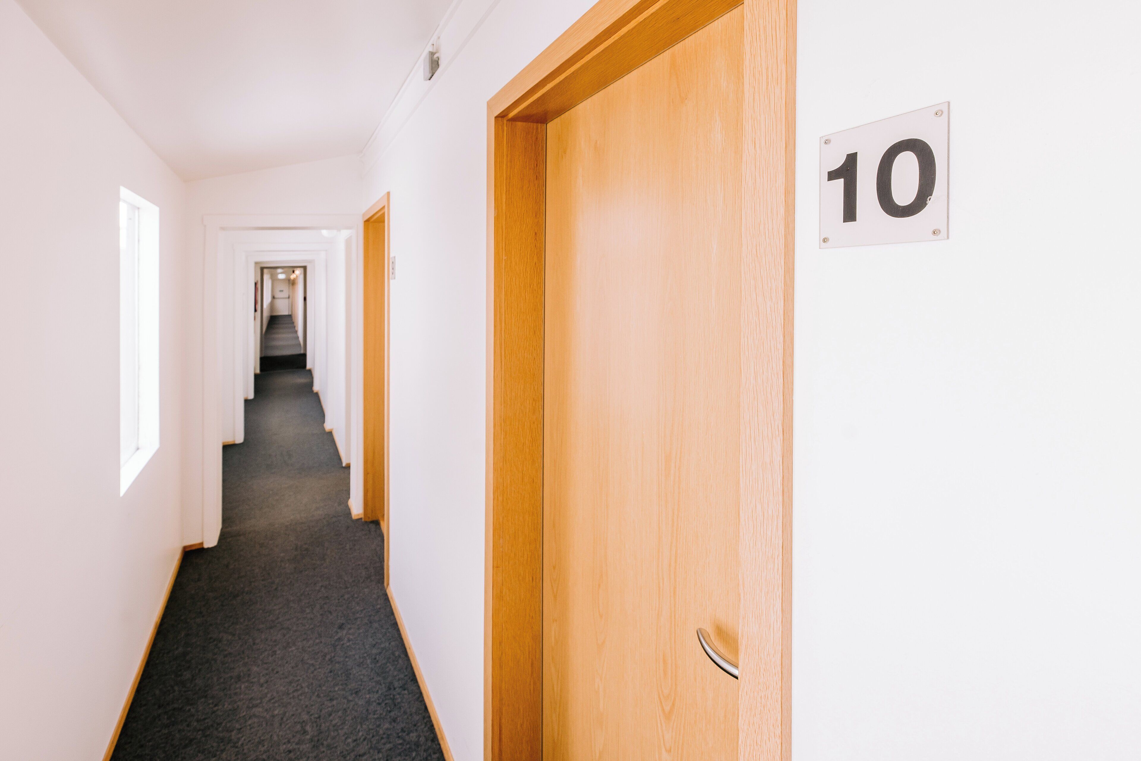 A hallway with a carpeted floor, a wooden door with a number sign, and a staircase in the distance.
