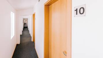 A hallway with a carpeted floor, a wooden door with a number sign, and a staircase in the distance.