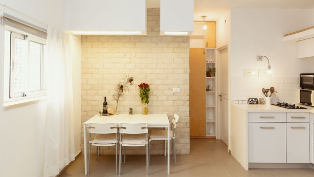 A compact kitchen with white cabinetry, a dining table with chairs, and a brick accent wall. - 2Rooms Perfect Location  1Min to  Beach (Tel Aviv)