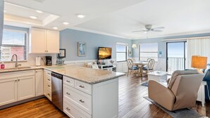 A modern kitchen with white cabinets, a dishwasher, and a granite countertop. There is a dining area with a table and chairs, and a ceiling fan.