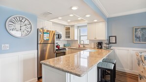 A modern kitchen with white cabinets, a stainless steel refrigerator, a granite countertop, and a large wall clock.