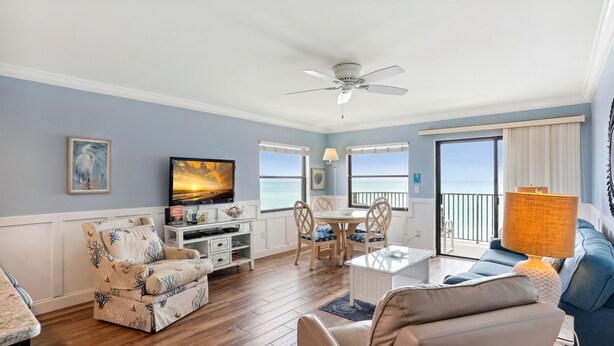 A living room with a TV, seating, a dining area, and a balcony with a view of the sea.