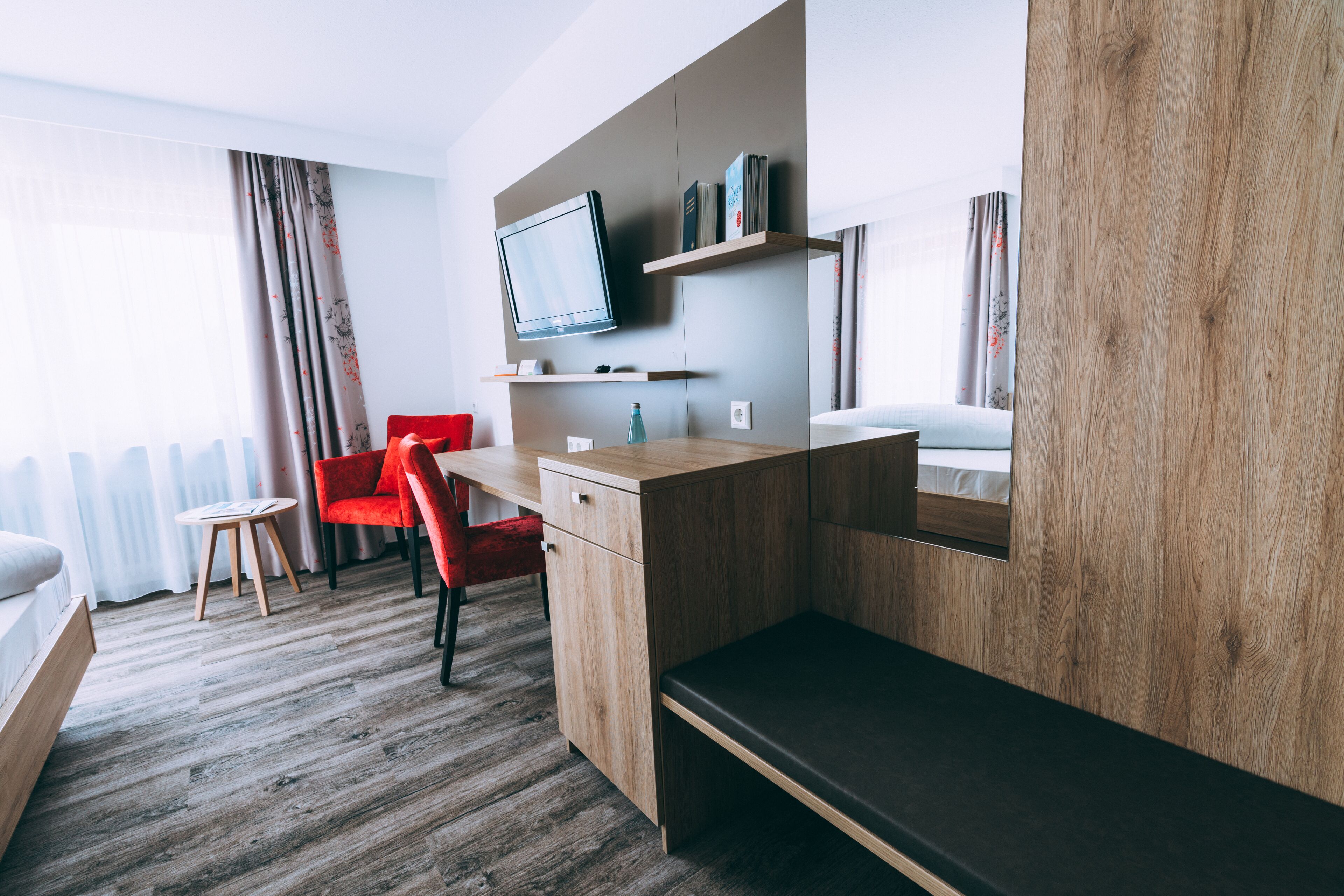 Une chambre d’hôtel moderne avec un bureau en bois, une chaise rouge, un lit et un téléviseur fixé au mur.