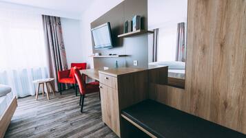 A modern hotel room with a wooden desk, a red chair, a bed, and a television mounted on the wall.