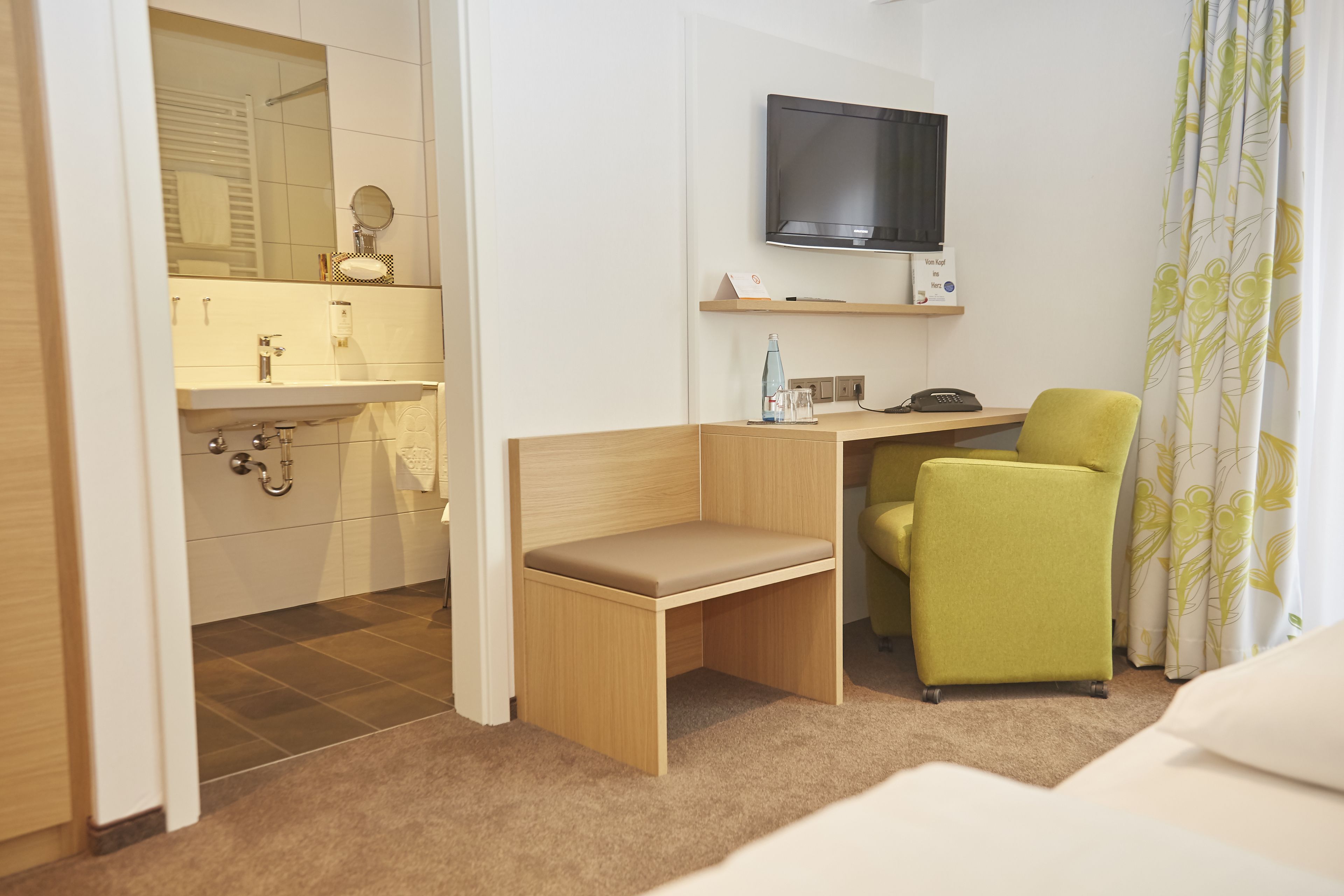 Habitación de hotel compacta con escritorio de madera, una silla verde, televisor y baño con espejo y lavabo.
