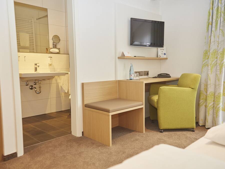 Habitación de hotel compacta con escritorio de madera, una silla verde, televisor y baño con espejo y lavabo.