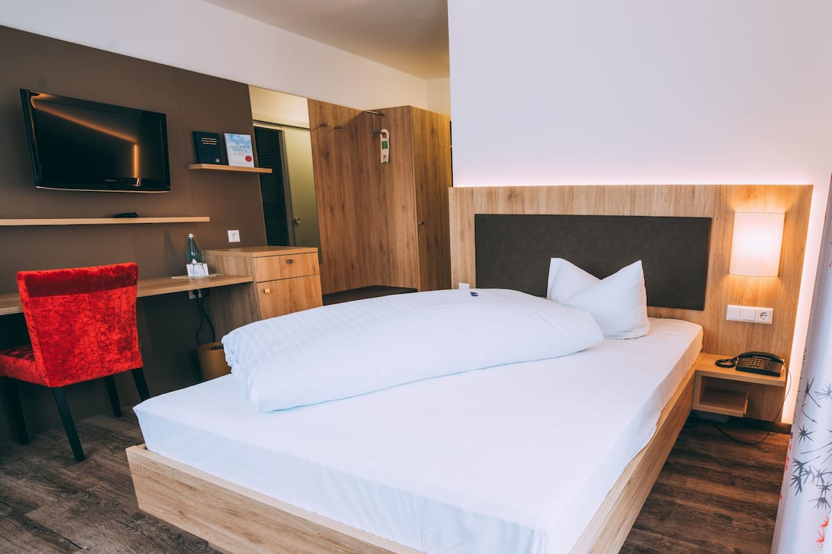 A hotel room with a large bed, a desk with a chair, a television, and a bedside table with a lamp.
