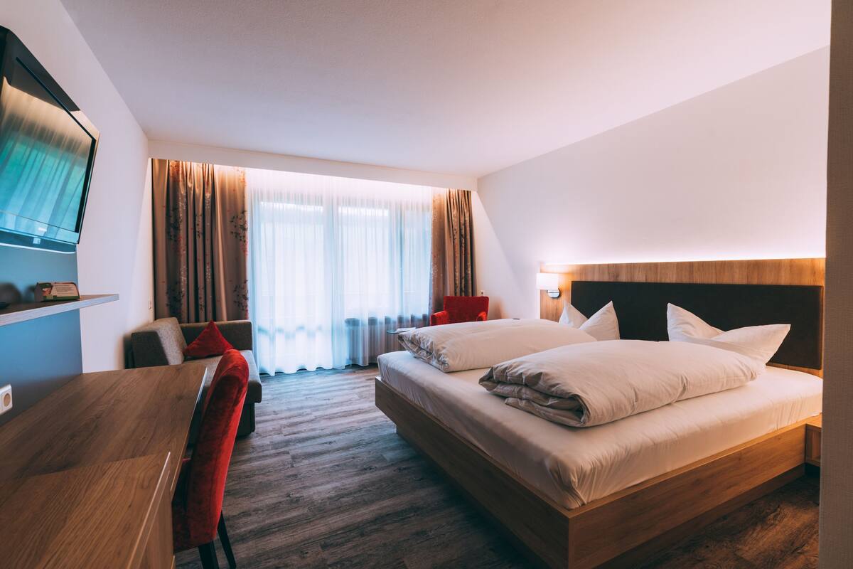 A hotel room with a wooden bed, a desk, a red chair, and a TV mounted on the wall.