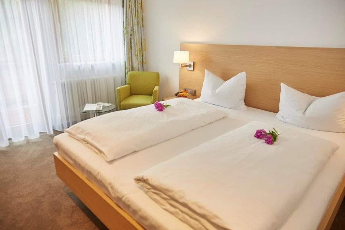 A hotel room with two beds, a small round table, a green armchair, and a window with sheer curtains.