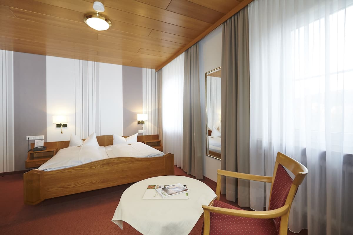 A hotel room with a wooden bed, a round table with a magazine, a chair, and a large mirror.