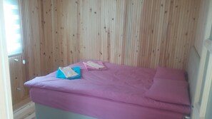 Une pièce avec un mur lambrissé en bois, un lit recouvert d’une courtepointe violette et une fenêtre avec des stores.