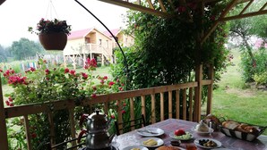 Une table dressée pour un repas, avec différents plats, une théière et un panier de pain, sur une terrasse couverte d’où l’on peut admirer une maison en bois et de la verdure.