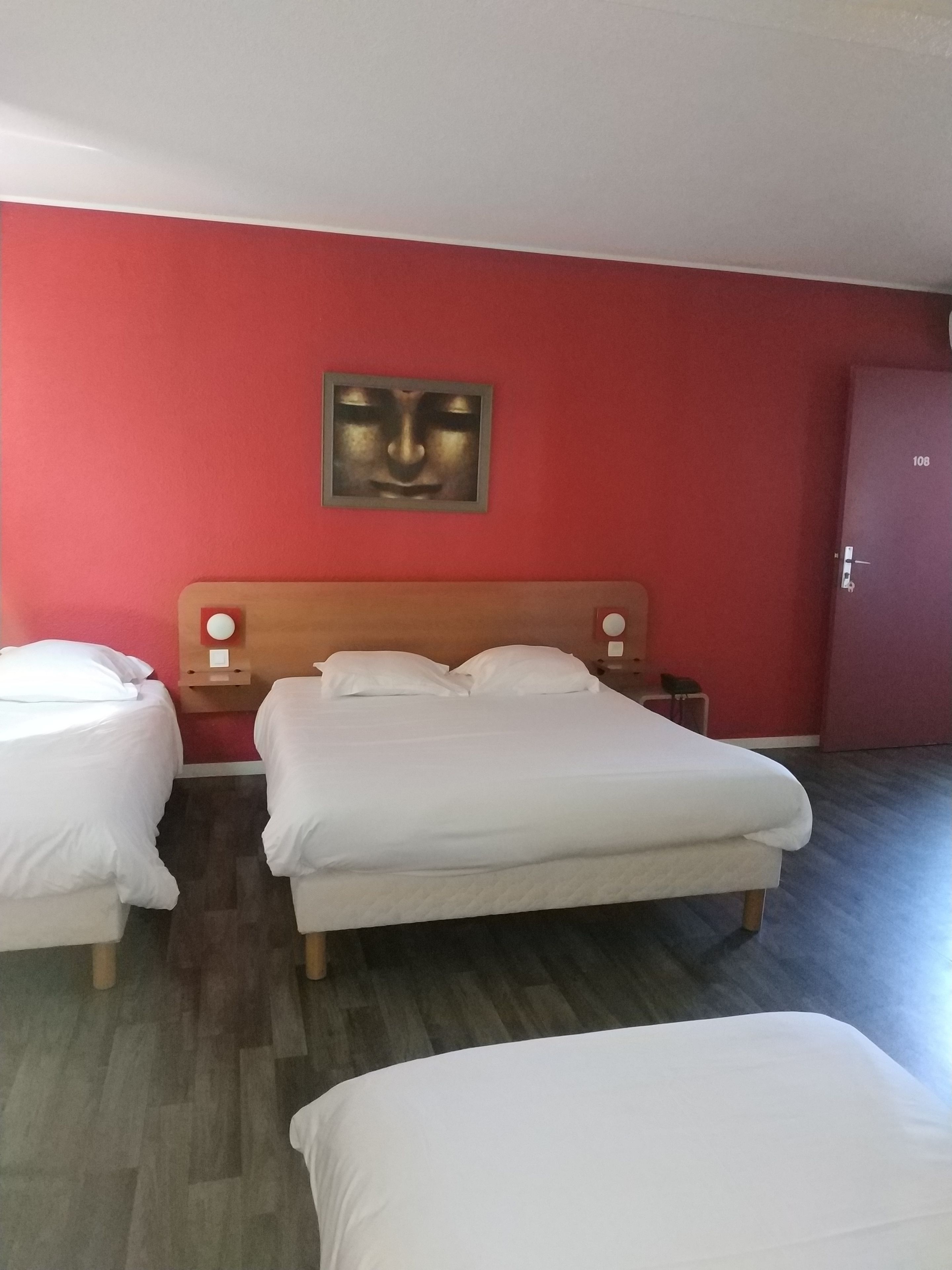 Une chambre d’hôtel avec un grand lit, deux tables de chevet, une petite table et un mur rouge orné d’une œuvre d’art encadrée.