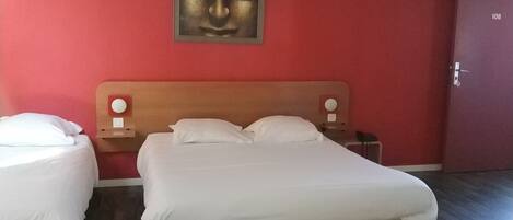 Une chambre d’hôtel avec un grand lit, deux tables de chevet, une petite table et un mur rouge orné d’une œuvre d’art encadrée.