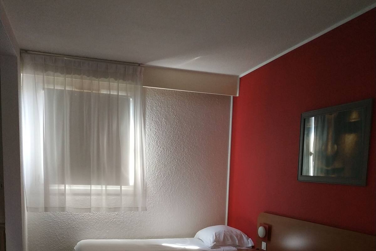 A hotel room with two beds, a red accent wall, a window with sheer curtains, and a painting on the wall.