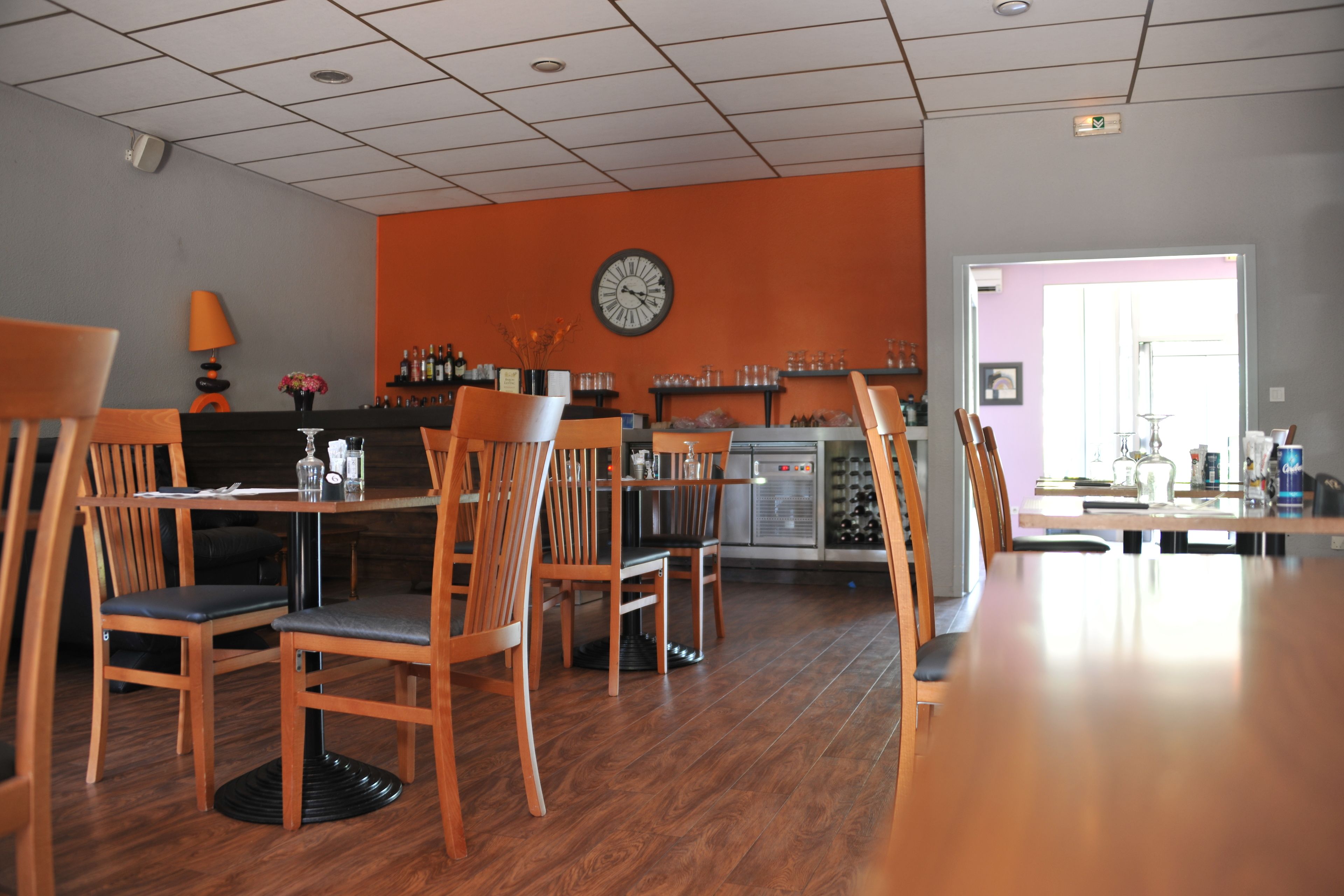 A restaurant with wooden chairs and tables, a clock on the wall, and a bar area with bottles and glasses.