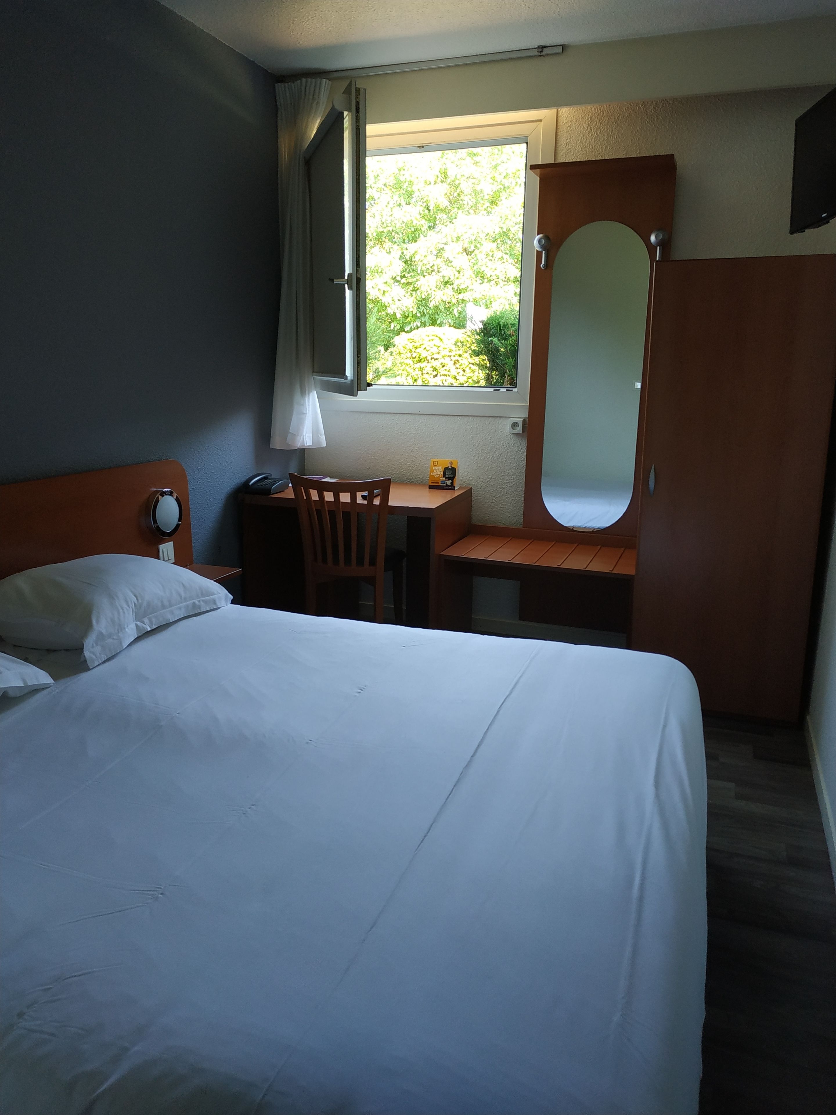 Habitación de hotel con cama, escritorio, silla, espejo y ventana con vistas a zonas verdes.