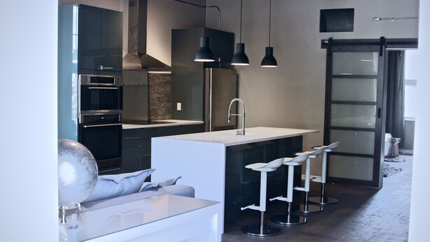 Una cocina moderna con armarios oscuros, electrodomésticos de acero inoxidable y encimera blanca. Hay tres taburetes de bar y lámparas colgantes sobre la encimera.