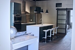 Una cocina moderna con armarios oscuros, electrodomésticos de acero inoxidable y encimera blanca. Hay tres taburetes de bar y lámparas colgantes sobre la encimera.