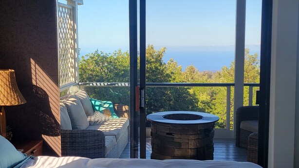 A bedroom with a large bed, a nightstand, a lamp, and a view of a balcony with outdoor furniture and a fire pit. - Big Private Mt. Home Minutes to Volcanos National Park
Myra's Blue Hawaii House  (Ocean View)